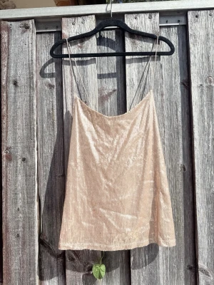 Beige glittrig linnetopp med tunna band - Snygg beige linnetopp med glittrigt tyg och tunna axelband. Toppen har en rak passform och är perfekt för dig som gillar en stilren men ändå festlig look. Materialet känns lätt och luftigt, perfekt för varma dagar.