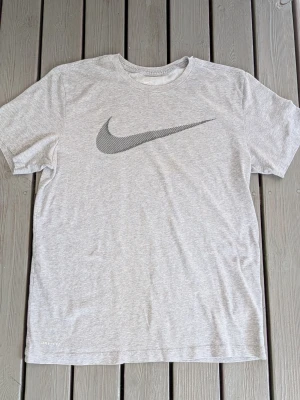 Grå Nike t-shirt med stor logga - Snygg grå t-shirt från Nike med stor svart swoosh-logga på bröstet. Klassisk rund halsringning och korta ärmar. Tillverkad i mjuk bomull som känns skön mot huden. Perfekt för en avslappnad och sportig stil.