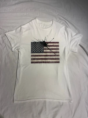 Vit t-shirt med amerikansk flagga - Vit t-shirt med tryck av amerikanska flaggan och svart färgstänk över bröstet. Klassisk passform och rund halsringning. Perfekt för dig som vill ha en statement-tee med unik twist. Tshirten är helt ny. Storlek: M/L | Pris: 340 SEK.