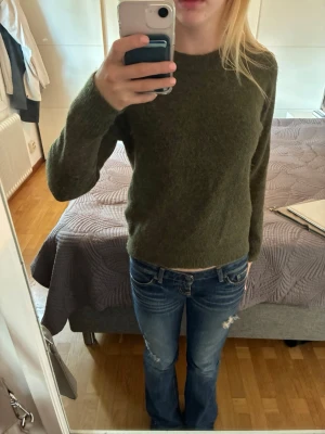 Olivgrön stickad tröja - Mysig olivgrön stickad tröja med rund halsringning och långa ärmar. Tröjan har en enkel, klassisk design och är gjord i ett mjukt material som känns skönt mot huden. Perfekt att bära till jeans eller kjol för en avslappnad look.