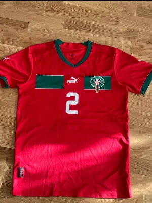Marocko fotbollströja Puma röd - Säljer en röd Marocko fotbollströja från Puma med gröna detaljer och nummer 2 på bröstet. Tröjan har grönt kantband vid halsen och ärmarna samt Marockos fotbollsförbunds märke på bröstet. Perfekt för dig som älskar fotboll och vill stötta Marocko!