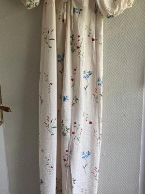 Lång blommig klänning med puffärm - Vacker långklänning i ljus färg med blommigt mönster i blått, rött och grönt. Klänningen har puffärmar, v-ringning och knyt framtill. Perfekt för sommarens alla tillfällen.