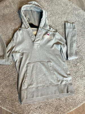  hoodie från Hollister - Snygg grå hoodie från Hollister med knappdetaljer vid halsen och magficka framtill. Klassisk logga och fågelbrodyr på bröstet. Tillverkad i mjukt bomullsmaterial och har en skön huva. Perfekt för en avslappnad och trendig look.