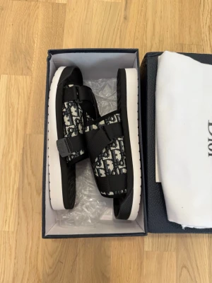 Alpha Svarta sandaler  - Snygga svarta sandaler  med vita och svarta monogramdetaljer på remmarna. Sandalerna har breda textilband och en vit, räfflad sula för extra komfort. Perfekta för dig som vill ha en trendig och lyxig look till sommaren.