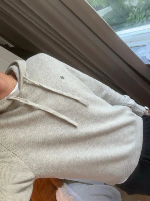 Beige Morris hoddie  - Säljer en stilren ljusgrå hoodie med huva och dragsko. Hoodien har ett diskret broderat emblem på bröstet och ribbade muddar vid ärmar och nederkant. Perfekt för en avslappnad look och passar till det mesta. Materialet känns mjukt och bekvämt.
