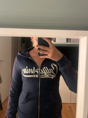 Mörkblå hoodie med tryck - Snygg mörkblå hoodie med dragkedja och vit text 'California' på bröstet. Har huva, fickor framtill och röda detaljer längs ärmarna. Perfekt för en avslappnad och sportig stil.💗