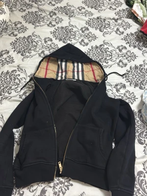 Svart hoodie med mönstrad huva - Snygg svart hoodie med dragkedja och fickor framtill. Huvan har ett klassiskt rutigt mönster på insidan i beige, rött och svart. Perfekt för en chill och stilren look. Dragsko i huvan för justerbar passform.