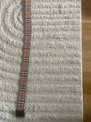 Rutig beige bälte med svart spänne - Snyggt bälte i klassiskt rutigt mönster med beige, rött och svart. Bältet har ett svart spänne med initialer och passar perfekt för att lyfta vilken outfit som helst. Materialet känns slätt och stilrent.
