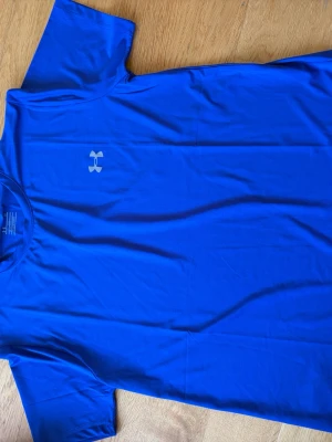 Blå tränings-tshirt från Under Armour - Snygg blå tränings-tshirt från Under Armour i funktionsmaterial. T-shirten har korta ärmar, rund halsringning och en diskret logga på bröstet. Perfekt för gymmet eller löprundan.