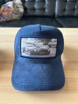 Mörkblå truckerkeps med bilmotiv - Snygg mörkblå truckerkeps med mesh baktill och justerbar snapback. Framtill finns en patch med svartvitt motiv av en klassisk sportbil. Kepsen har en mjuk, nästan frottéliknande textur på framsidan och skärmen.