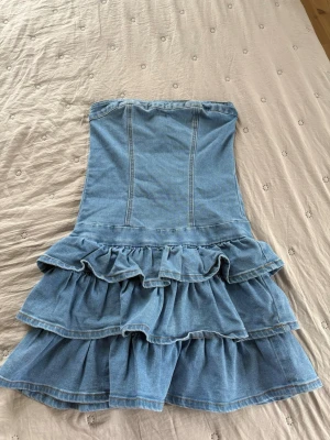 Axelbandslös jeansklänning med volanger - Säljer en snygg axelbandslös jeansklänning med flera lager volanger och dragkedja i ryggen. Klänningen har en kort modell och är perfekt för dig som gillar en trendig och lekfull stil. Tillverkad i klassiskt blått denimtyg.