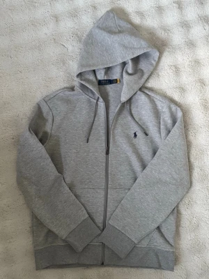 Grå zip hoodie ifrån Polo Ralph Lauren - Helt oanvänd Polo Ralph Lauren zip hoodie. Storlek L. Perfekt för dig som gillar en avslappnad men samtidigt trendig stil. Säljes pga för stor storlek. Tveka inte att höra av er ifall ni har frågor eller funderingar! 