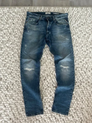Blå slitna jeans från Replay - Säljer ett par blå jeans från Replay med slitningar och distressed detaljer. Jeansen har fem fickor, knappgylf och klassisk passform. Perfekta för en avslappnad och trendig look.