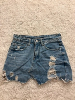 Blå jeanskjol med slitna detaljer - Kort blå jeanskjol i denim med slitna och trasiga detaljer samt fransig nederkant. Kjolen har klassisk femficksdesign, knapp och dragkedja framtill och råa kanter för en trendig och avslappnad stil.
