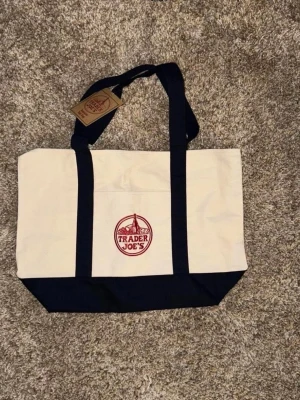 Trader Joe's canvas shoppingväska - Stilren shoppingväska i kraftig canvas från Trader Joe's. Väskan är vit med mörkblå botten och handtag samt har en röd logga framtill. Praktisk ytterficka och långa axelband gör den enkel att bära. Perfekt för shopping eller stranden.