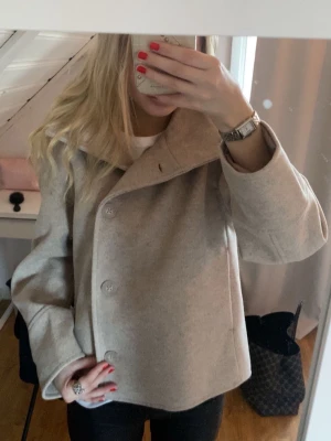 Beige kappa - Stilren beige kappa med knappar framtill och bred krage. Kortare modell som ger en modern look och passar perfekt till höst och vinter. 