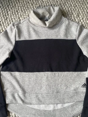 Stl S Grå polotröja med svart rand Adidas - Snygg grå polotröja från Adidas med bred svart rand över bröstet. Tröjan har lång ärm, hög krage och Adidas-logga nedtill. Tillverkad i mjukt sweatshirtmaterial, perfekt för chill eller träning. Finns en  lös tråd. Se bild 