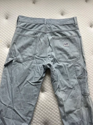 Leee randiga workwear jeans i modellen carpenter - Supersnygga jeans från märket Lee. Jeansen är i modellen carpenter och storlek W30 L32. Har flera snygga detaljer såsom ett band i sidan (bild 3) samt extra fickor i sidan (bild 4). Är i superfint skick, enda skavanker jag hittat är en liten fläck (bild 7) som smälter in med ränderna så den syns knappt!   Midjemått: 41cm Innerbenslängd: 75cm Grenhöjd: 32cm