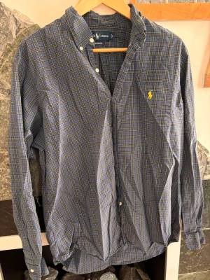 Blårutig skjorta från Ralph Lauren - Klassisk blårutig skjorta från Ralph Lauren i custom fit-modell. Skjortan har knappar framtill, lång ärm och den ikoniska gula loggan broderad på bröstet. Tillverkad i mjuk bomull och passar perfekt till jeans eller chinos.