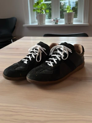 Svarta sneakers med vita snören -  BE OM MER BILDER VID INTRESSE!! Stilrena svarta sneakers med vita snören och bruna gummisulor. Skorna har en klassisk låg profil och är tillverkade i en mix av mocka och läder. Perfekta för en avslappnad och trendig look.