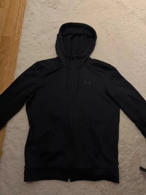 Svart hoodie från Under Armour - Snygg svart hoodie från Under Armour med dragkedja framtill och justerbar huva. Tillverkad i ett mjukt och lite stretchigt material som känns skönt mot huden. Diskret logga på bröstet och praktiska fickor på sidorna. Perfekt för en sportig och avslappnad stil.