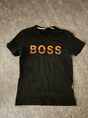 Svart BOSS t-shirt med orange tryck - Snygg svart t-shirt från BOSS med orange text på bröstet. Klassisk rund hals och korta ärmar. Tillverkad i mjuk bomull som känns skön mot huden. Perfekt för dig som vill ha en enkel men stilren look.
