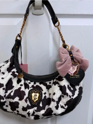 Juicy Couture axelväska med ko-mönster - Superfin axelväska från Juicy Couture med svartvitt ko-mönster, svarta läderdetaljer och guldfärgade kedjor. Väskan har en rosa rosett och hjärtberlock som dekoration samt ett emblem framtill. Perfekt för dig som vill sticka ut med din stil.