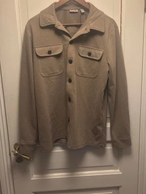 Beige overshirt från Only & Sons - Stilren beige overshirt från Only & Sons med klassisk krage, två bröstfickor med lock och svarta knappar framtill. Tillverkad i ett mjukt tyg som ger en avslappnad look. Perfekt att bära över en t-shirt för en trendig lager-på-lager stil. Ganska stor för sin storlek så sitter snarare som en Small. Overshirten är bra skick.
