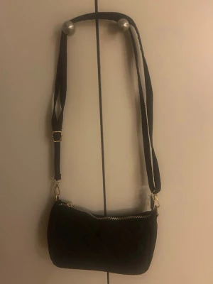 Svart quiltad axelväska - Snygg svart axelväska med quiltat mönster och justerbar axelrem. Väskan har guldfärgade detaljer och dragkedja upptill. Perfekt för dig som vill ha en stilren och trendig accessoar till din outfit.