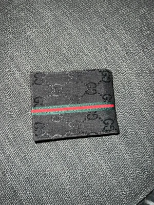Svart plånbok med GG-mönster - Snygg svart plånbok i tyg med klassiskt GG-mönster och en grön-röd rand tvärs över framsidan. Plånboken har en stilren och kompakt design, perfekt för kort och sedlar. Passar dig som gillar en clean och trendig look.