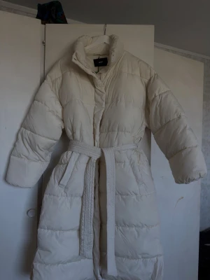 Lång beige dunjacka med bälte - Säljer en lång, beige dunjacka med hög krage och avtagbart midjebälte. Jackan har en quiltad design, två sidofickor och stängs med tryckknappar. Perfekt för kalla vinterdagar och ger en stilren look.