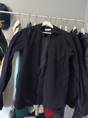 Mörkblå overshirt med dragkedja - Storlek 39 i fint skick. Manchester overshirt från John Henric.Stilren mörkblå overshirt med dragkedja framtill och klassisk krage. Overshirten har långa ärmar och är tillverkad i ett kraftigare tyg som ger en avslappnad men ändå snygg look. Perfekt att bära över en t-shirt eller hoodie.