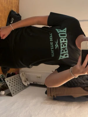 Svart Reebok t-shirt med tryck - Snygg svart t-shirt från Reebok med stort turkost tryck på bröstet och klassisk rund hals. Tillverkad i återvunnen polyester med Speedwick-teknologi som håller dig torr. Perfekt för dig som gillar sportig stil.