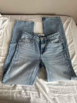 Ljusblå raka jeans från Pull&Bear - Säljer ett par ljusblå jeans från Pull and Bear med rak passform och klassisk femficksdesign. Jeansen har hög midja, gylf med dragkedja och knapp samt subtila slitningar för en avslappnad look. Perfekta till en casual outfit.