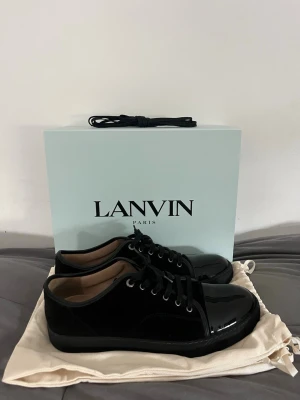 Lanvin cap toe  - Snygga svarta sneakers från Lanvin med blank lackad tå och ovandel i mocka. Skorna har svarta skosnören, metallöljetter och en klassisk låg silhuett. Perfekta för dig som vill ha en stilren och exklusiv look med en lyxig touch. Nyskick 
