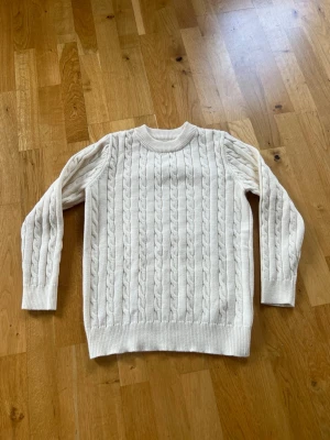Vit kabelstickad tröja - 150 cl, Kabelstickad tröja i varm vit ton med rund halsringning och ribbade muddar. Klassisk design med långa ärmar och grovstickat mönster som ger en tidlös look. Perfekt för kyliga dagar och enkel att matcha med olika outfits.Nyskick, bara provad.