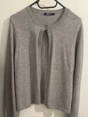 Grå kofta från Gina Tricot - Stilren grå kofta från Gina Tricot med rund halsringning och en knapp framtill. Koftan har långa ärmar och ribbade kanter vid ärmslut och nederkant. Perfekt att slänga över en topp för en enkel och snygg look.