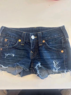 Jeans shorts True Religion  - Säljer ett par blå jeansshorts med råa, fransiga kanter och slitna detaljer från True Religion. Shortsen har klassiska fem fickor, synliga sömmar och knappgylf. Bakfickan har en tydlig dekorationssöm. Perfekta för sommaren!