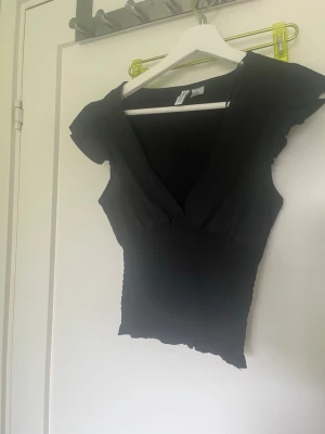 Svart v-ringad topp med volangärm - Säljer en svart topp med v-ringning och volangärmar. Toppen är smockad i midjan för en snygg passform och har en lätt croppad känsla. Perfekt att styla med jeans eller kjol för en trendig look.