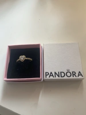 Guldfärgad ring med hjärta från Pandora - Supersöt guldfärgad ring från Pandora med ett hjärtformat motiv i mitten, dekorerad med gnistrande stenar runt hjärtat och längs bandet. Kommer i original ask. Perfekt för dig som gillar eleganta och romantiska smycken.