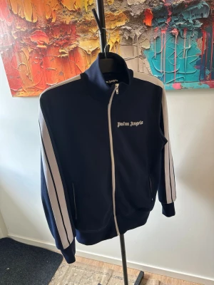 Palm Angels marinblå ziptröja - Snygg marinblå ziptröja från Palm Angels med vita ränder längs ärmarna och logga på bröstet. Tröjan har hög krage, dragkedja framtill och ribbade muddar. Perfekt för en sportig och trendig look.
