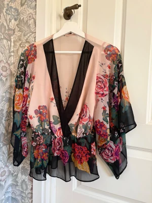 Blommig transparent blus med v-ringning - Säljer en superfin blus i transparent material med stora färgglada blommor på ljusrosa botten. Blusen har djup v-ringning med svarta detaljer och vida, trekvartsärmar. Perfekt för dig som gillar romantisk och bohemisk stil.