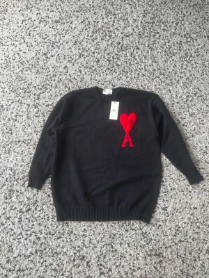 Svart stickad ami Paris tröja med rött hjärta - Snygg svart stickad tröja med ett stort rött hjärta och bokstaven A broderat på bröstet. Tröjan har rund halsringning, långa ärmar och ribbade muddar vid ärmslut och nederkant. Perfekt för dig som vill ha en trendig och stilren look.