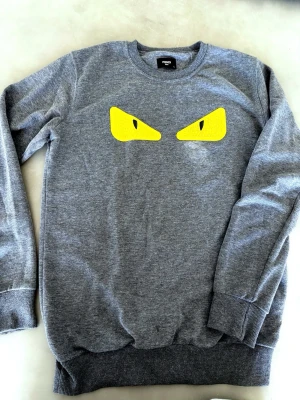 Fendi tröja grå - Cool grå sweatshirt med ett par stora gula ögon på bröstet. Tröjan har rund halsringning, ribbade muddar vid ärmslut och nederkant samt en avslappnad passform. Perfekt för dig som vill sticka ut med en unik detalj.
