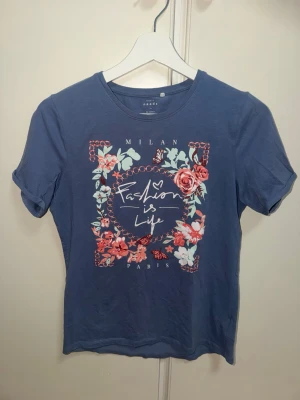 Blå t-shirt med blommigt tryck - Snygg blå t-shirt med blommigt tryck och texten 'Fashion is Life' framtill. T-shirten har rund halsringning och korta ärmar. Perfekt för dig som gillar färgglada detaljer och vill sticka ut lite extra.