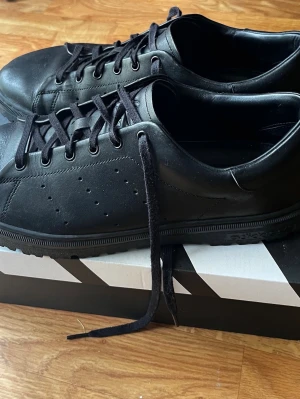 Svarta Adidas lädersneakers - Klassiska svarta sneakers från Adidas i läder med svarta skosnören och diskreta perforerade detaljer på sidorna. Låg modell med stilren design och gummisula. Perfekta för dig som gillar en clean och tidlös look.