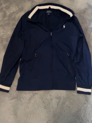 Mörkblå polo ralph lauren jacka - Jag säljer en mörkblå polo Ralph lauren regnjacka som knappt har används jag har haft på mig den 3-4 gånger och den är nästan helt ny utan några skador nypriset är cirka 2400kr den går att använda året runt fast under vintern kan det bli lite kyligt men annars kan man ha på den året runt