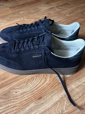Svarta sneakers i mocka från Gant - Stilrena svarta sneakers från Gant i mjuk mocka med mörkbruna gummisulor. Klassisk låg modell med svarta skosnören och diskret Gant-logga i guld på sidan. Insidan är ljus och skon har en tidlös, clean look som passar till det mesta.