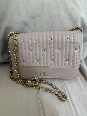 Ljusrosa axelväska från Bebe - Snygg ljusrosa axelväska från Bebe med guldfärgad kedjerem och quiltad design med små nitar. Väskan har ett elegant metallogo framtill och är tillverkad i konstläder. Perfekt för dig som vill ha en trendig och stilren accessoar.