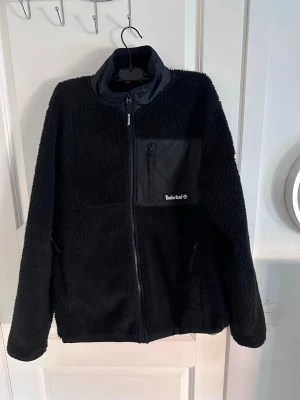 Svart teddyjacka från Timberland - Mysig svart teddyjacka från Timberland med hög krage och dragkedja framtill. Jackan har en bröstficka i nylon med dragkedja och Timberland-logga, samt två sidofickor. Perfekt för kalla dagar och ger en cool streetstil. Använd fåtal gånger, storlek M